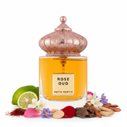 Frasco de perfume Rose Oud de Matin Martin, botella rectangular transparente con líquido ámbar dorado y etiqueta dorada, tapón cúpula morisca acanalada en oro rosa envejecido con remate esférico, sobre fondo blanco, rodeado de azafrán, bergamota, rosa, geranio, jazmín, madera de oud, sándalo y cedro.