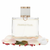 Frasco Prestige Pearl EDP, botella rectangular redondeada en cristal transparente con líquido crema pálido, texto dorado PRESTIGE PEARL, perla blanca lústrosa en bezal oro rosa en el hombro, banda dorada en la base y tapa rectangular blanca mate con banda dorada, sobre fondo blanco, rodeado de pimienta rosa, pétalos de rosa, pachulí y almizcle.