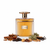 Frasco Riqqa de Ard Al Zaafaran EDP, botella rectangular redondeada con acanalado vertical profundo en vidrio transparente con líquido ámbar-coñac, banda de texto dorado en hombros, placa dorada con monograma árabe plateado y texto RIQQA, anillo dorado en cuello y gran tapa disco circular plana dorada, sobre fondo blanco, rodeado de coñac, canela, haba tonka, musgo de roble, praliné, vainilla y sándalo.