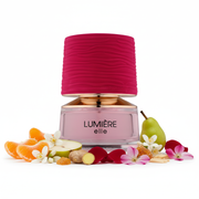 Frasco de perfume Lumière Elle de Maison Alhambra EDP, botella rectangular redondeada transparente con líquido rosa blush pálido y texto negro LUMIÈRE elle impreso en el vidrio, collar ancho oro rosa pulido, gran tapa cilíndrica envuelta en tela magenta-fucsia con textura acanalada horizontal, sobre fondo blanco, rodeado de mandarina, pera, jengibre, jazmín, azahar, rosa, ámbar gris, sándalo y flor peruana.